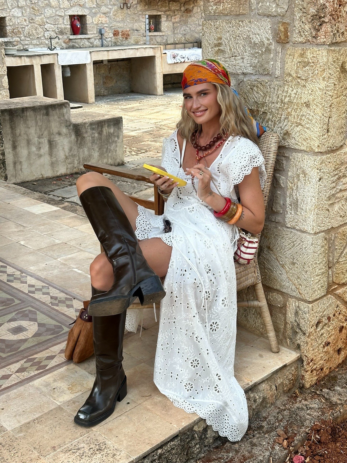 Modelo senta relaxada com vestido branco de algodão bordado, botas pretas de couro, lenço colorido na cabeça e acessórios