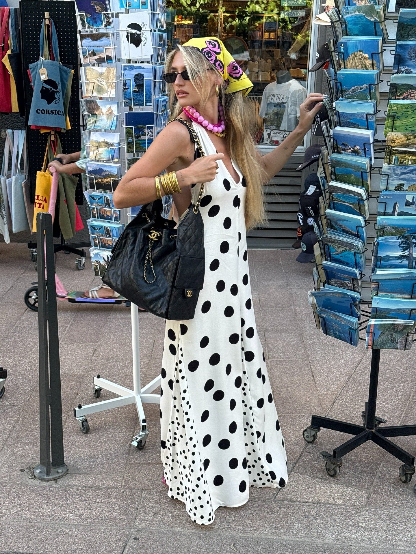 Modelo posa com vestido longo branco de poás pretos em tecido leve, lenço amarelo com flores, acessórios vibrantes e bolsa