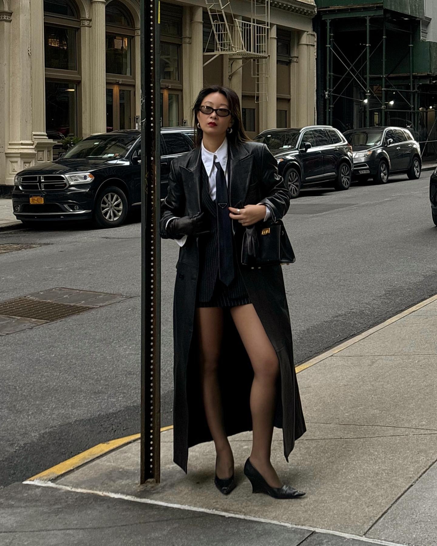 Modelo posa na rua com scarpin preto de bico fino, meia-calça, blazer oversized, camisa branca e sobretudo longo de couro.