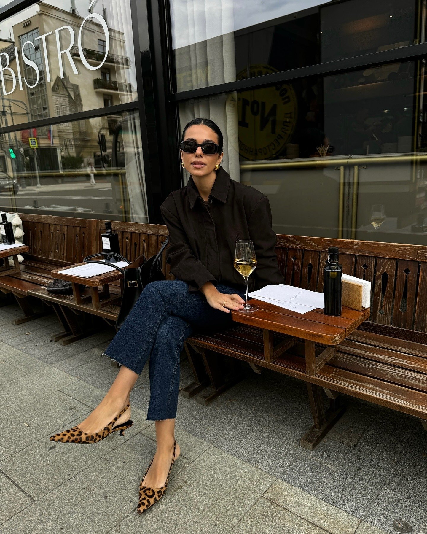 Modelo posa sentada em bistrô urbano com scarpin de salto baixo e bico fino em estampa animal print, combinando jaqueta