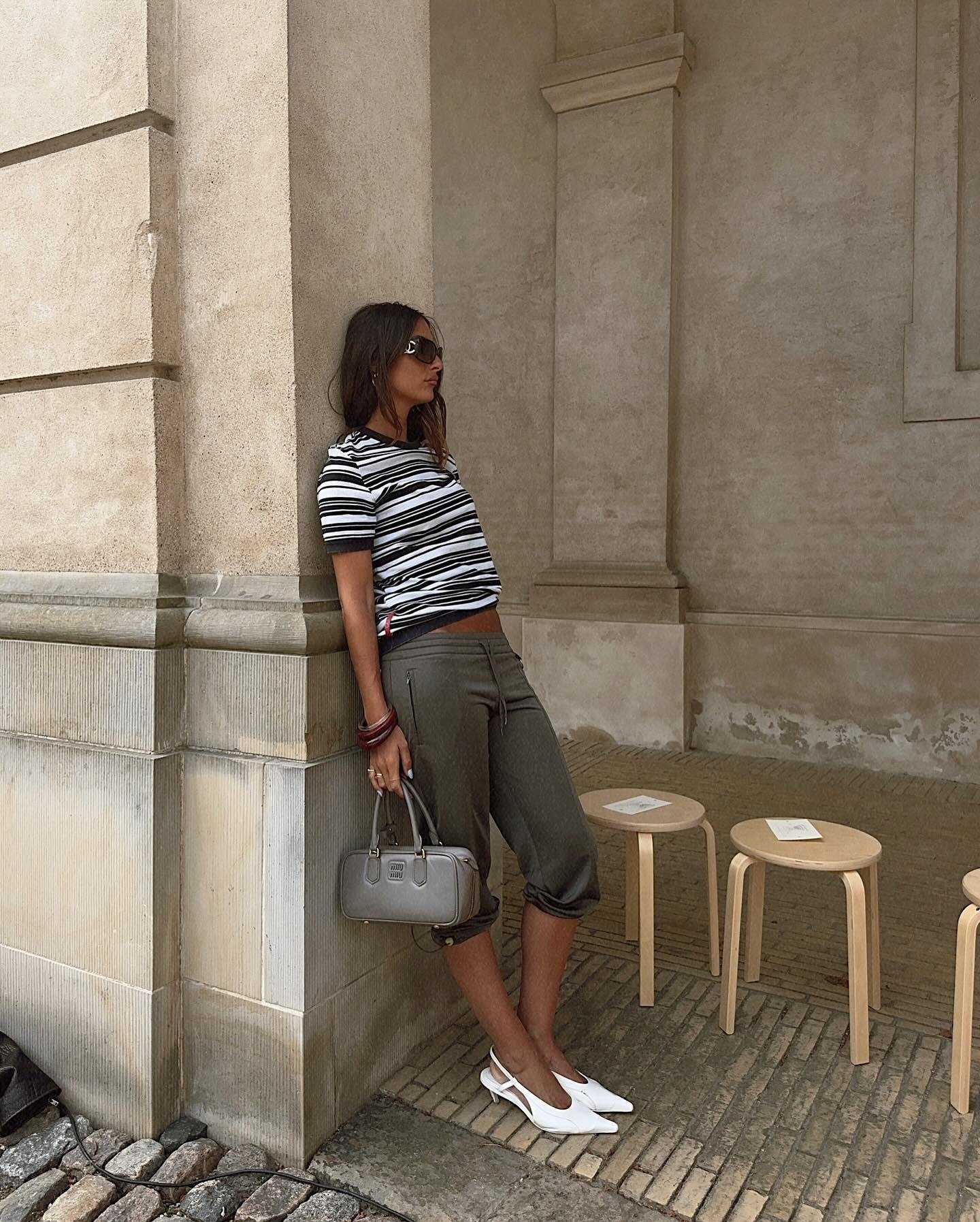 Modelo posa encostada em parede com scarpin branco de bico fino e salto baixo, combinando com calça capri cinza e blusa