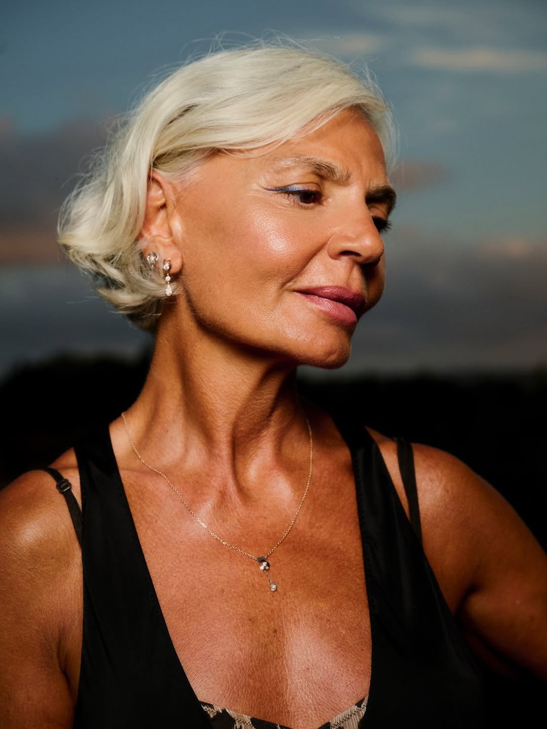 Mulher madura com pele radiante e base luminosa, cabelo branco curto, vestido preto e joias delicadas em clima sereno.