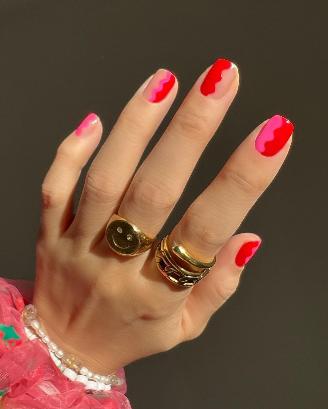 Unhas para Natal 2025 com design ondulado em vermelho e rosa vibrante, combinando anéis dourados e pulseira de pérolas.