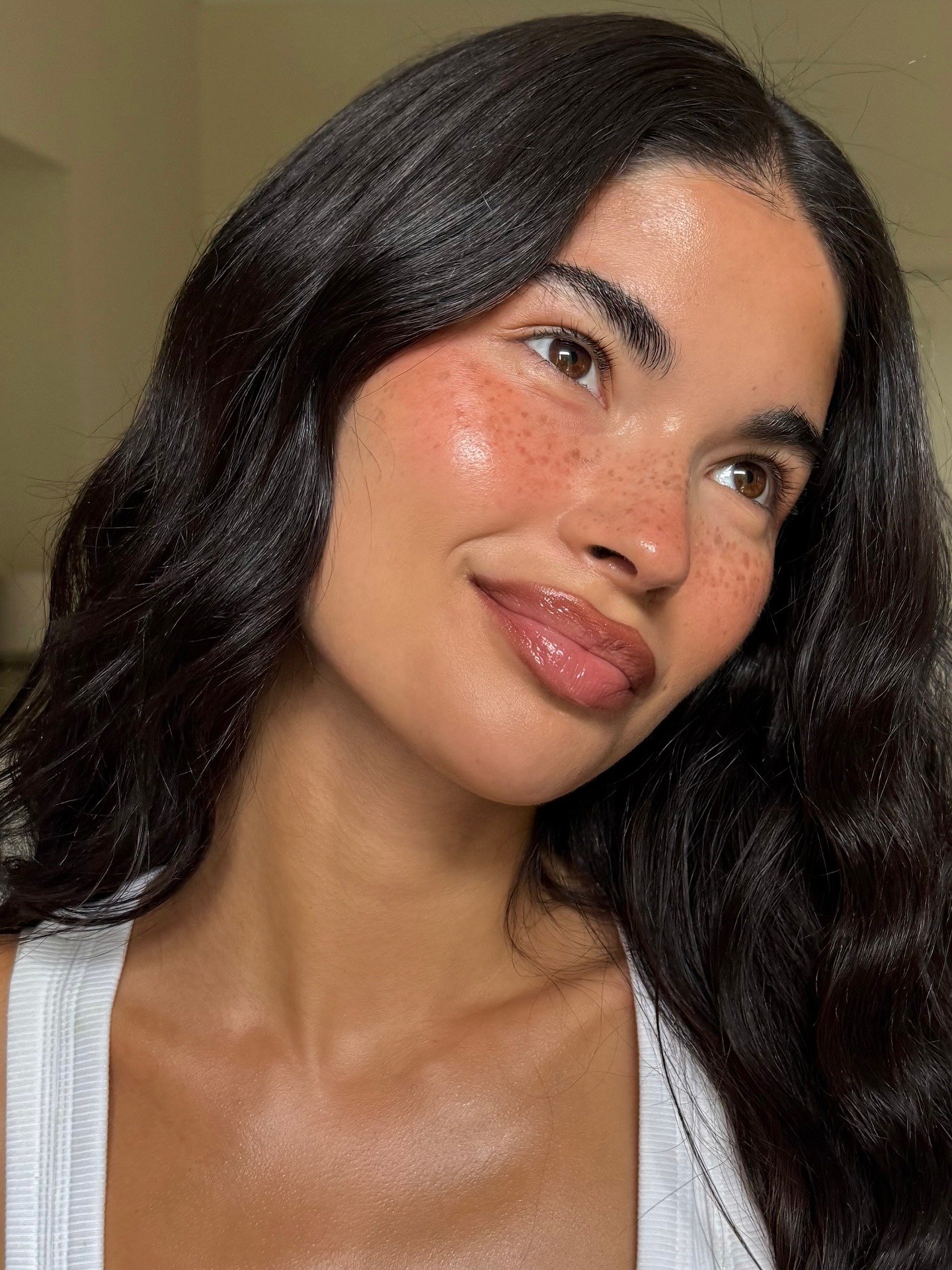 Modelo com pele luminosa e natural, lábios com brilho suave e cabelo ondulado preto, evidenciando fixadores de maquiagem