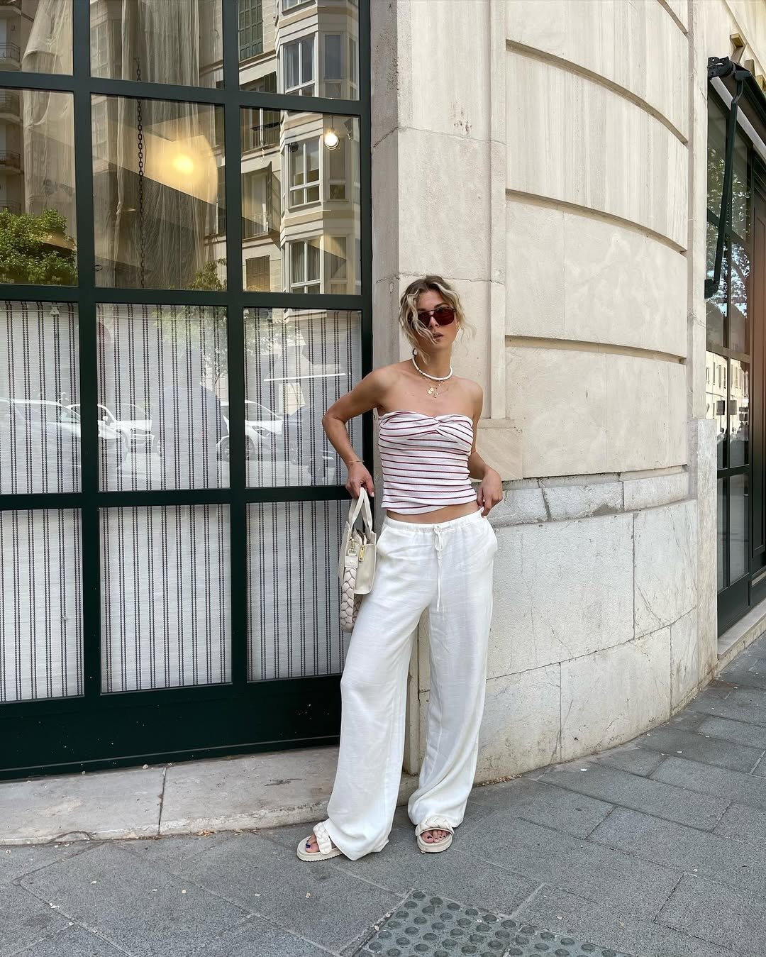 Modelo posa encostada em parede urbana com top tomara que caia listrado, calça branca ampla e papete branca trançada.