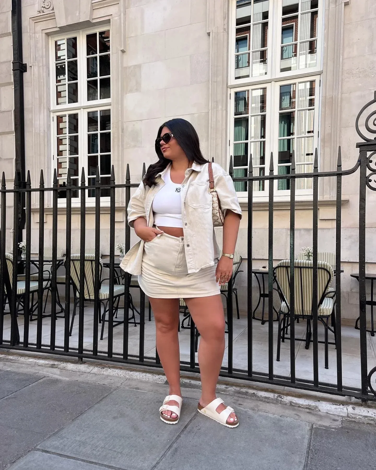 Modelo posa com papete branca de tiras largas em look casual com saia e camisa oversized bege, transmitindo conforto urbano.