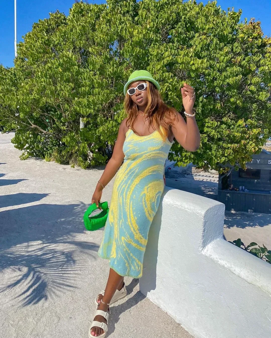 Modelo posa com vestido midi azul e amarelo de tricô, papete branca de tiras largas, chapéu verde e bolsa de mão felpuda