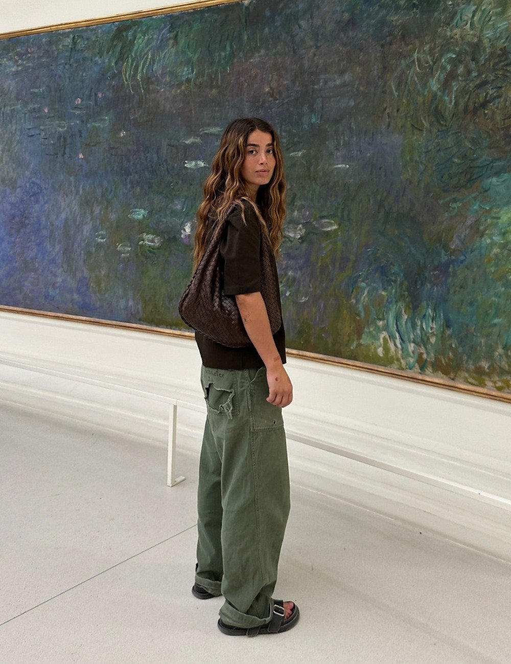 Modelo posa de perfil com papete feminina preta de tiras largas, calça cargo verde larga, camiseta preta e bolsa marrom