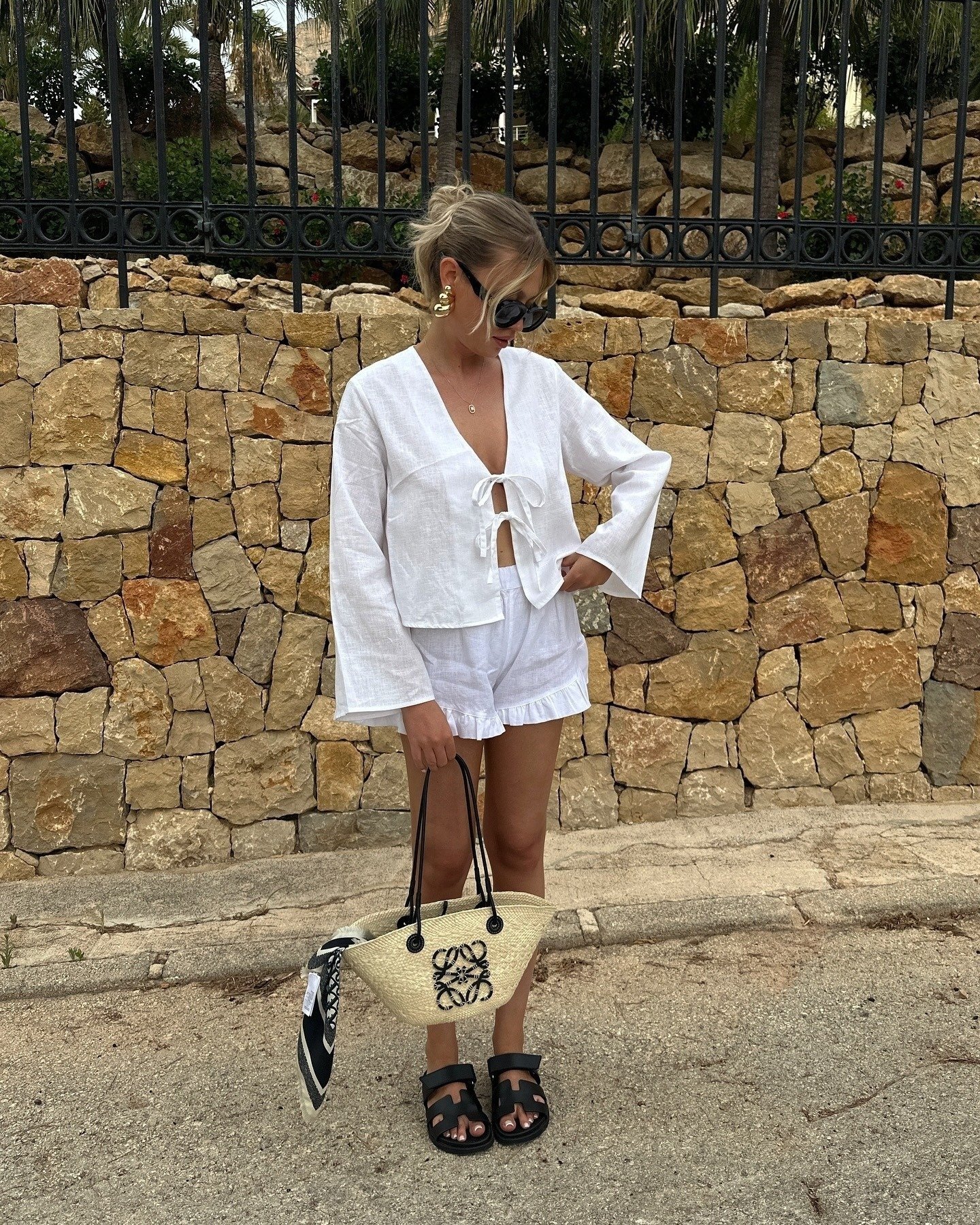 Modelo posa com look branco de linho fluido, shorts com babados e papete feminina preta de tiras largas, em clima casual e