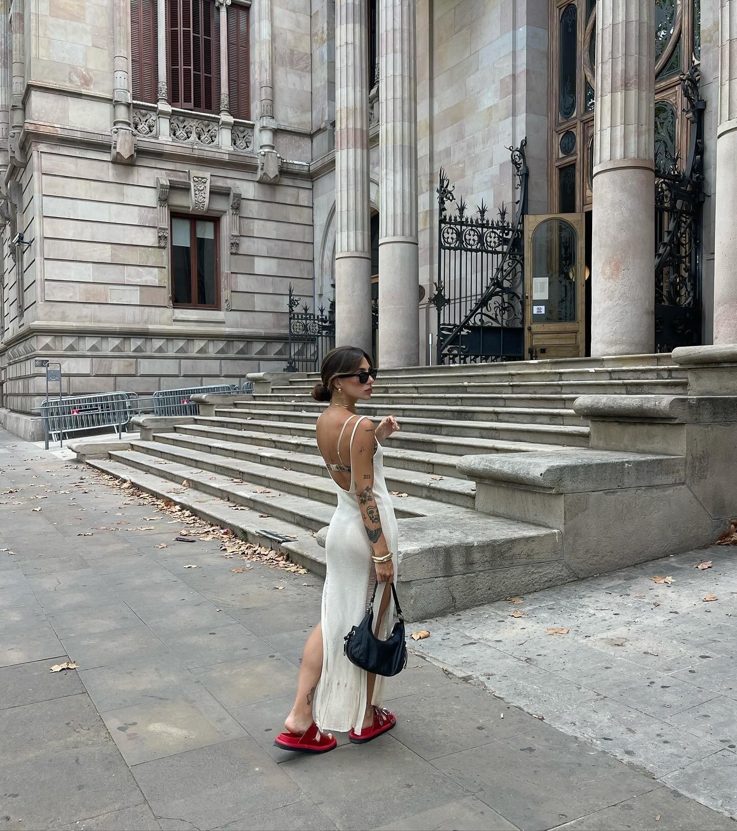 Modelo posa de costas em ambiente urbano usando vestido branco longo justo de alças finas e papete feminina vermelha com