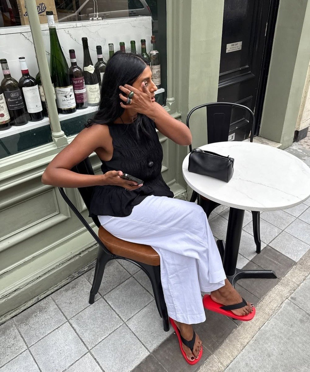 Mulher sentada em cadeira preta com calça branca de linho ampla, regata preta texturizada e chinelos vermelhos, em clima