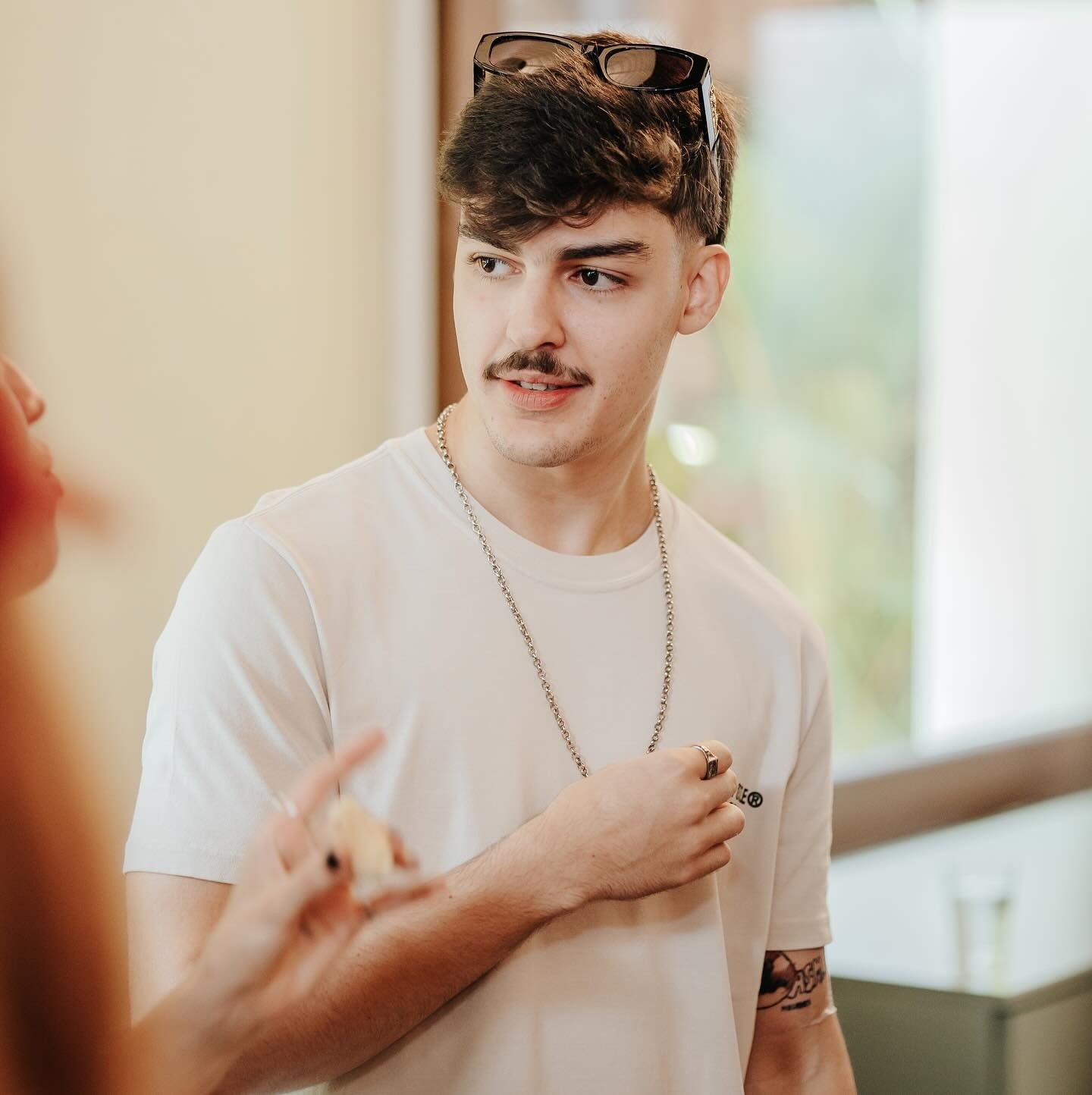 Homem com camiseta branca, corrente e anel prata, posa conversando em ambiente claro, sugerindo estilo casual para perfumes