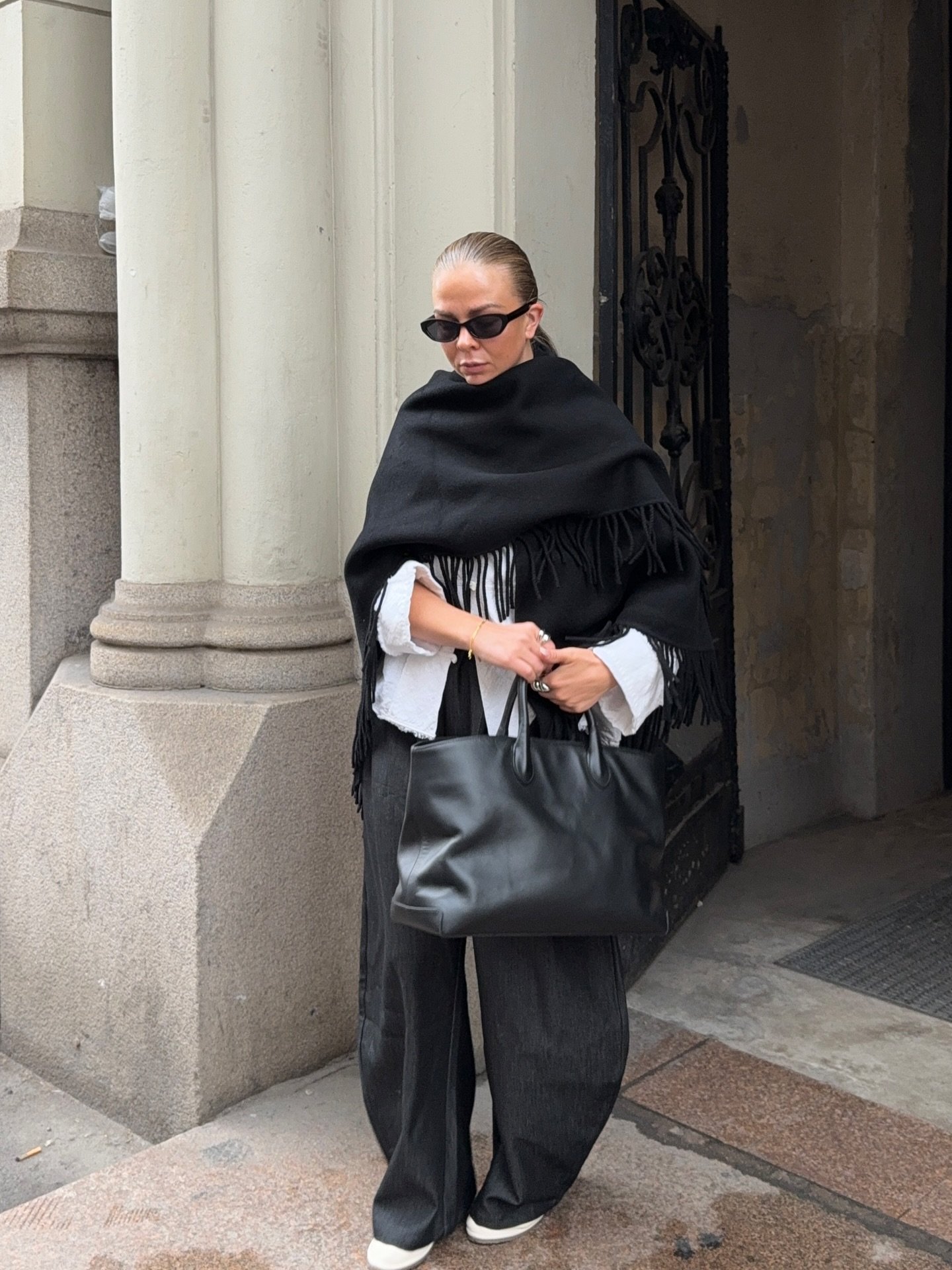 Modelo posa com poncho preto de lã com franjas, combinando com camisa branca, calça larga preta e bolsa de couro, em clima