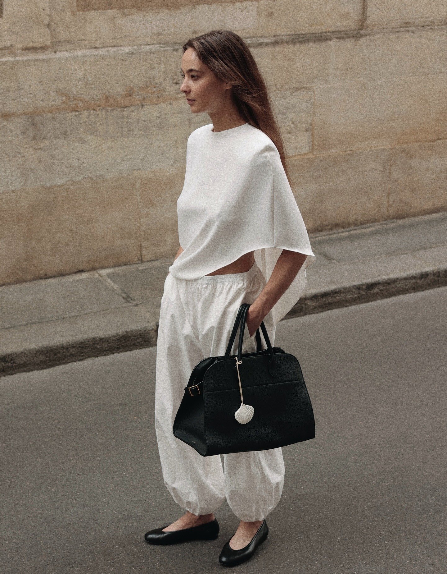 Modelo caminha na rua com poncho branco de tecido fluido e modelagem solta, combinando com calça ampla branca e bolsa preta