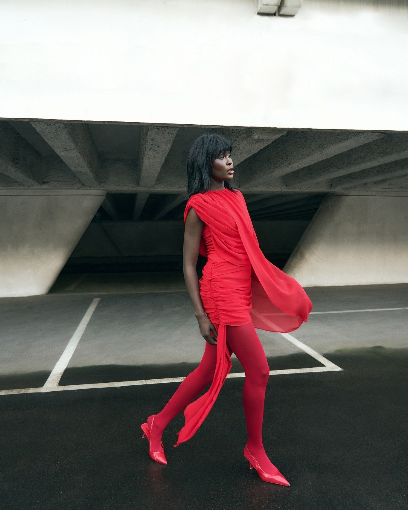 Modelo caminha com postura confiante usando poncho vermelho fluido de tecido leve sobre vestido justo com drapeados .