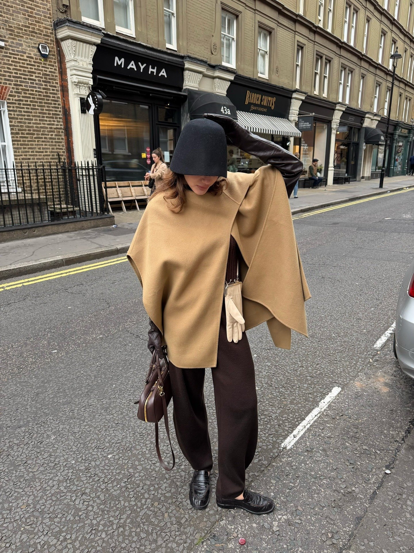 Modelo posa na rua usando poncho camel de tecido estruturado com calça marrom, luvas longas, chapéu preto e bolsa marrom