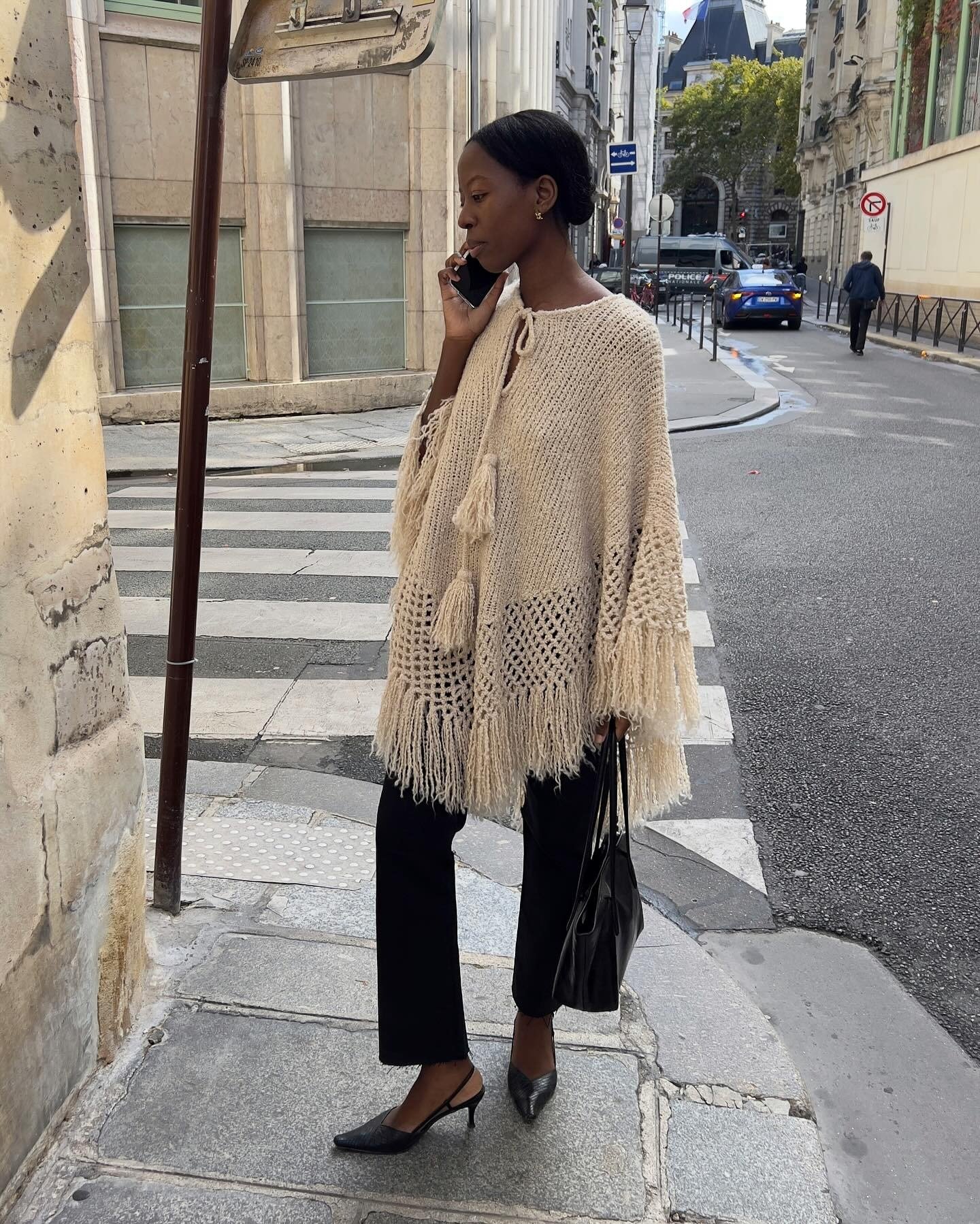 Modelo conversa ao celular usando poncho bege de tricô com franjas, calça preta reta e scarpin, em rua urbana sofisticada.