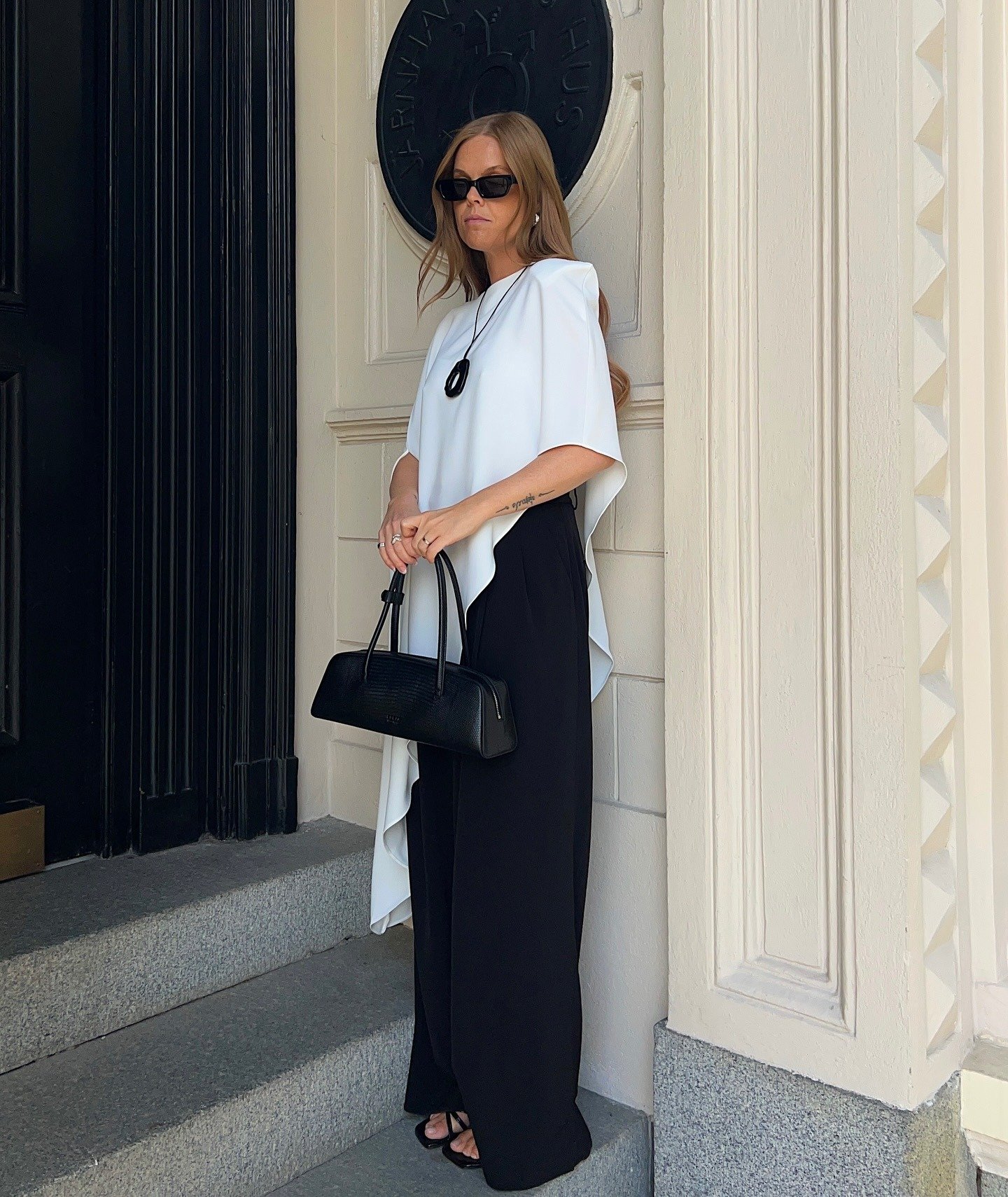 Modelo posa com poncho branco assimétrico de tecido fluido, combinando com calça preta ampla e acessórios minimalistas.