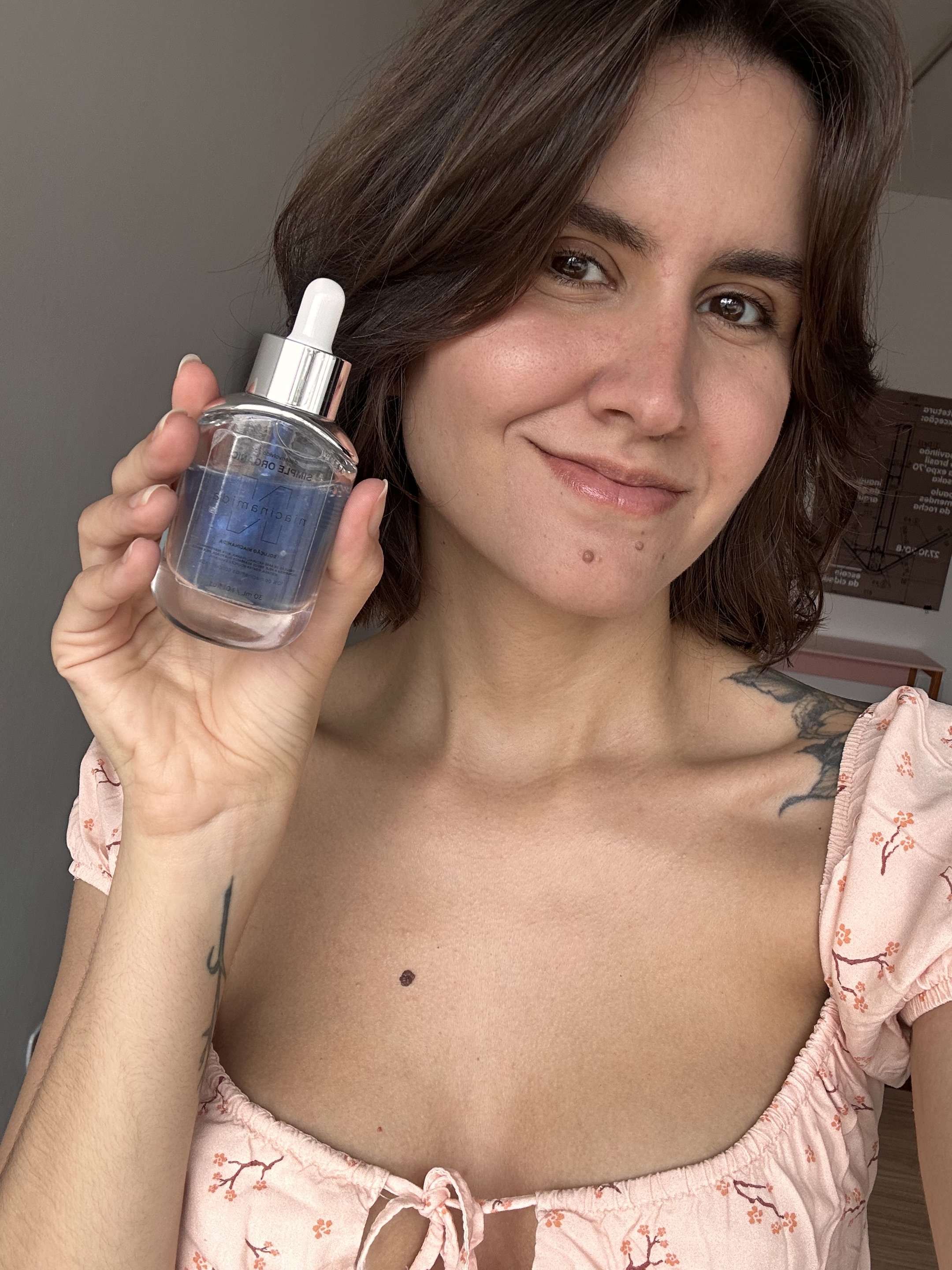 Modelo sorri segurando frasco de vidro com conta-gotas, veste blusa rosa clara depois de produtos para pele oleosa