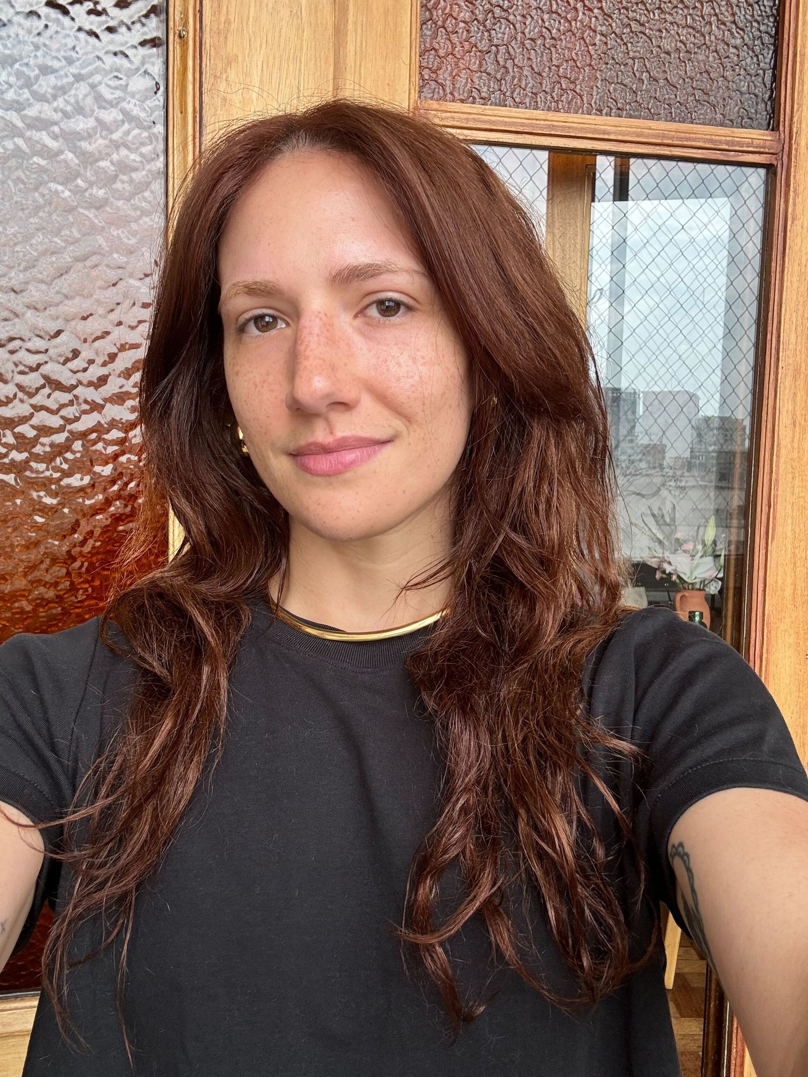 Mulher posa sorrindo com cabelo castanho ondulado solto, camiseta preta básica e colar dourado com produtos para pele oleosa