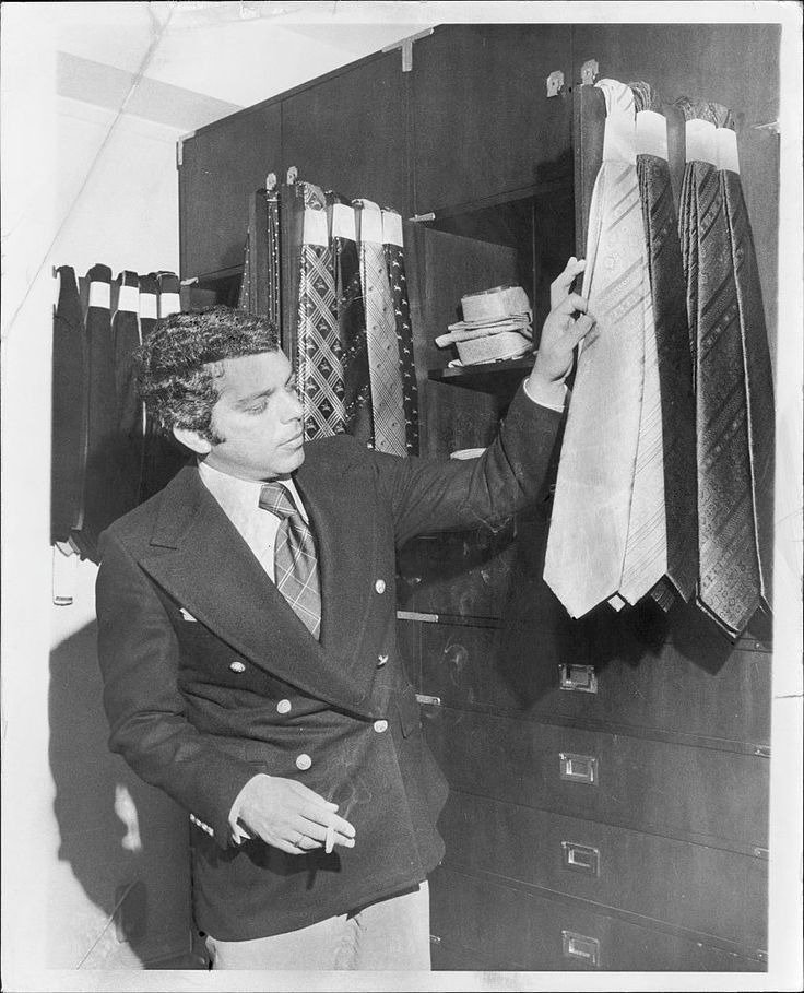 Homem com blazer duplo preto e gravata xadrez Ralph Lauren examina gravatas de seda em tons neutros, em ambiente clássico.