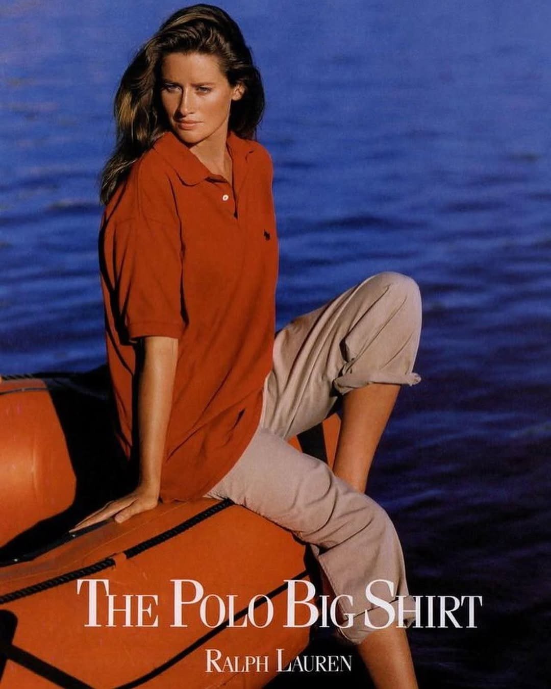 Modelo senta em bote laranja à beira d’água vestindo polo oversized terracota e calça bege dobrada, Ralph lauren