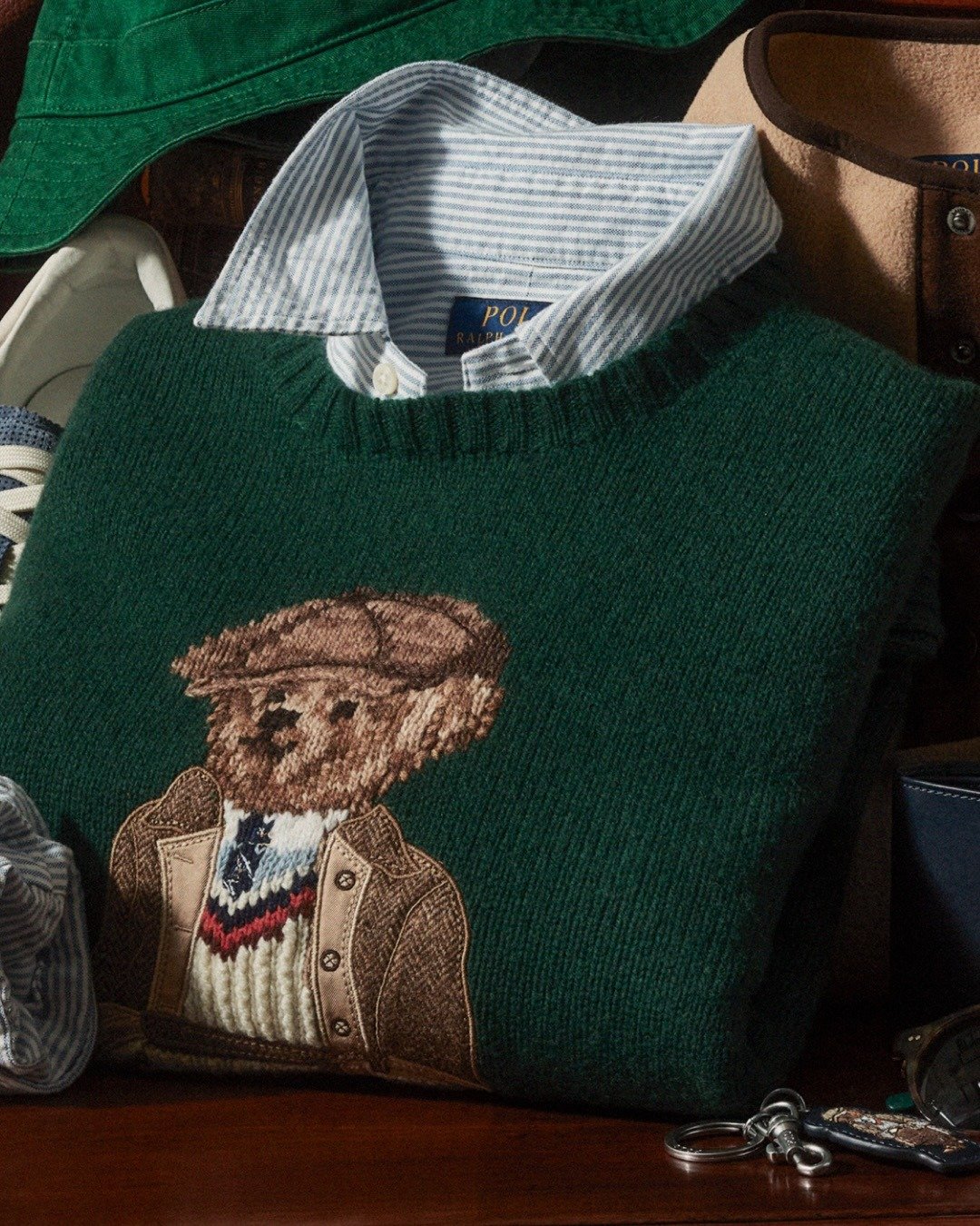 Suéter verde de lã Ralph Lauren com bordado de urso estilizado, camisa azul listrada e acessórios em tom terroso.
