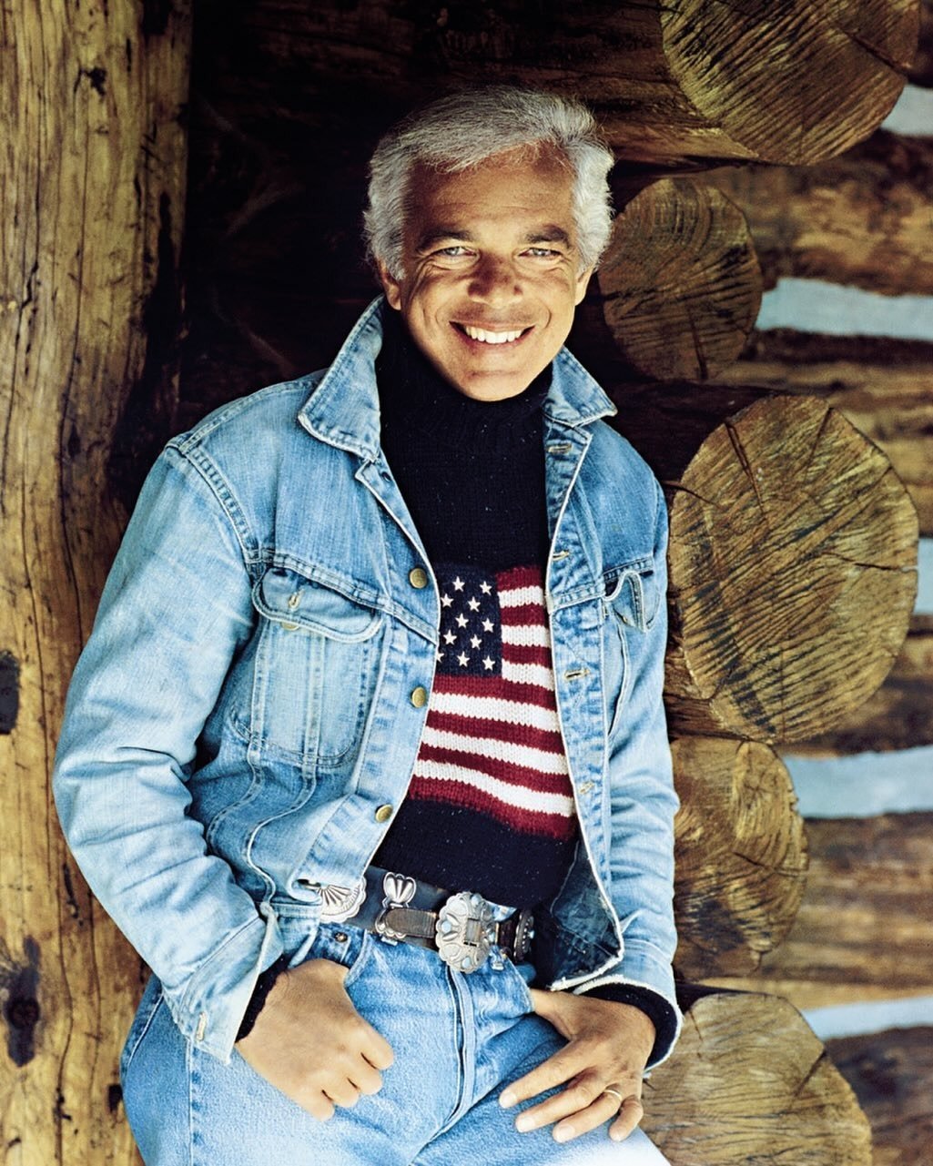 Ralph Lauren sorri e posa com jaqueta jeans clara, suéter preto com bandeira dos EUA em tricô e cinto de fivela prata.