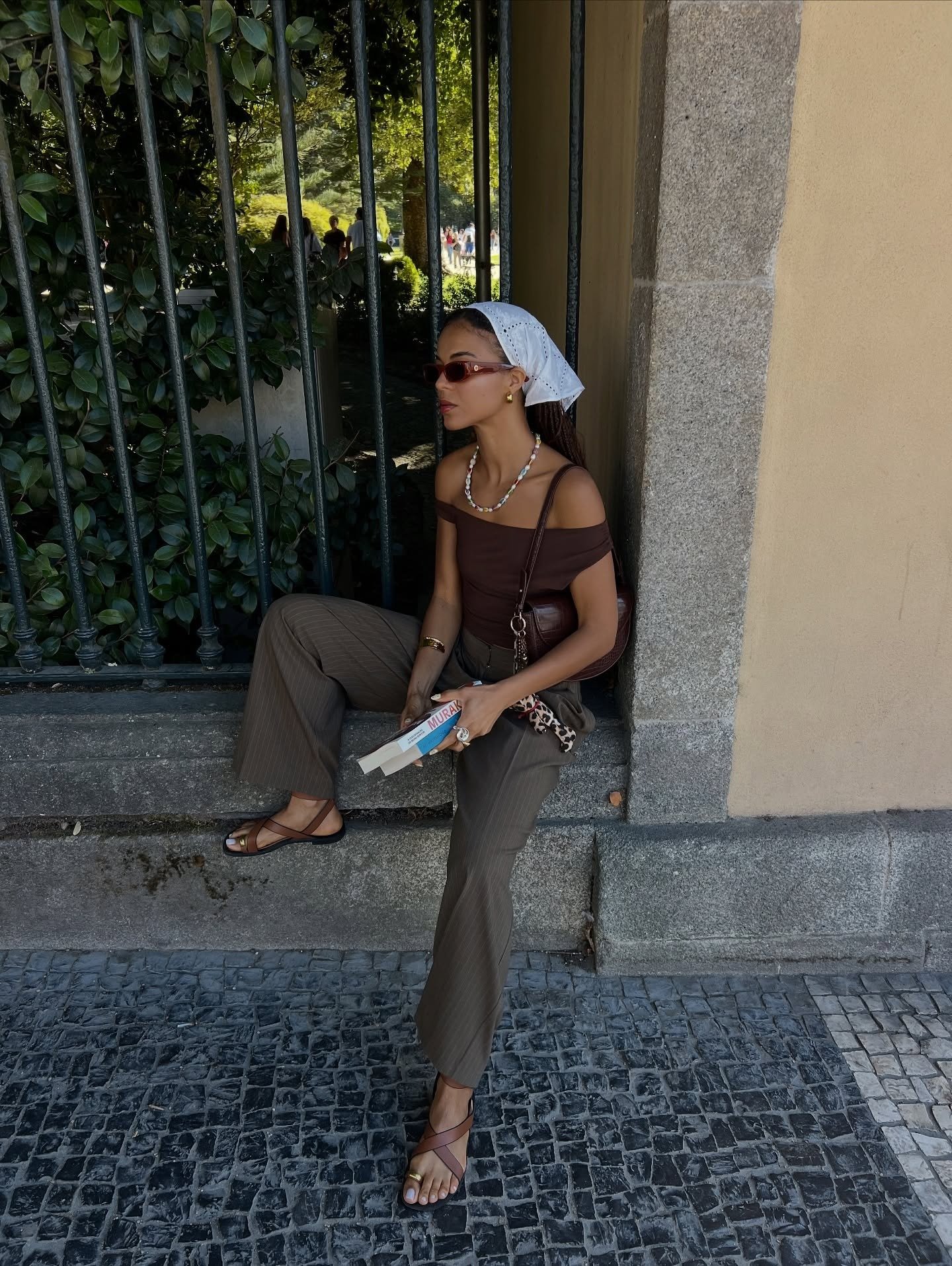 Modelo senta casualmente com postura relaxada usando modelos de rasteirinha em tiras finas marrom, combinando com calça