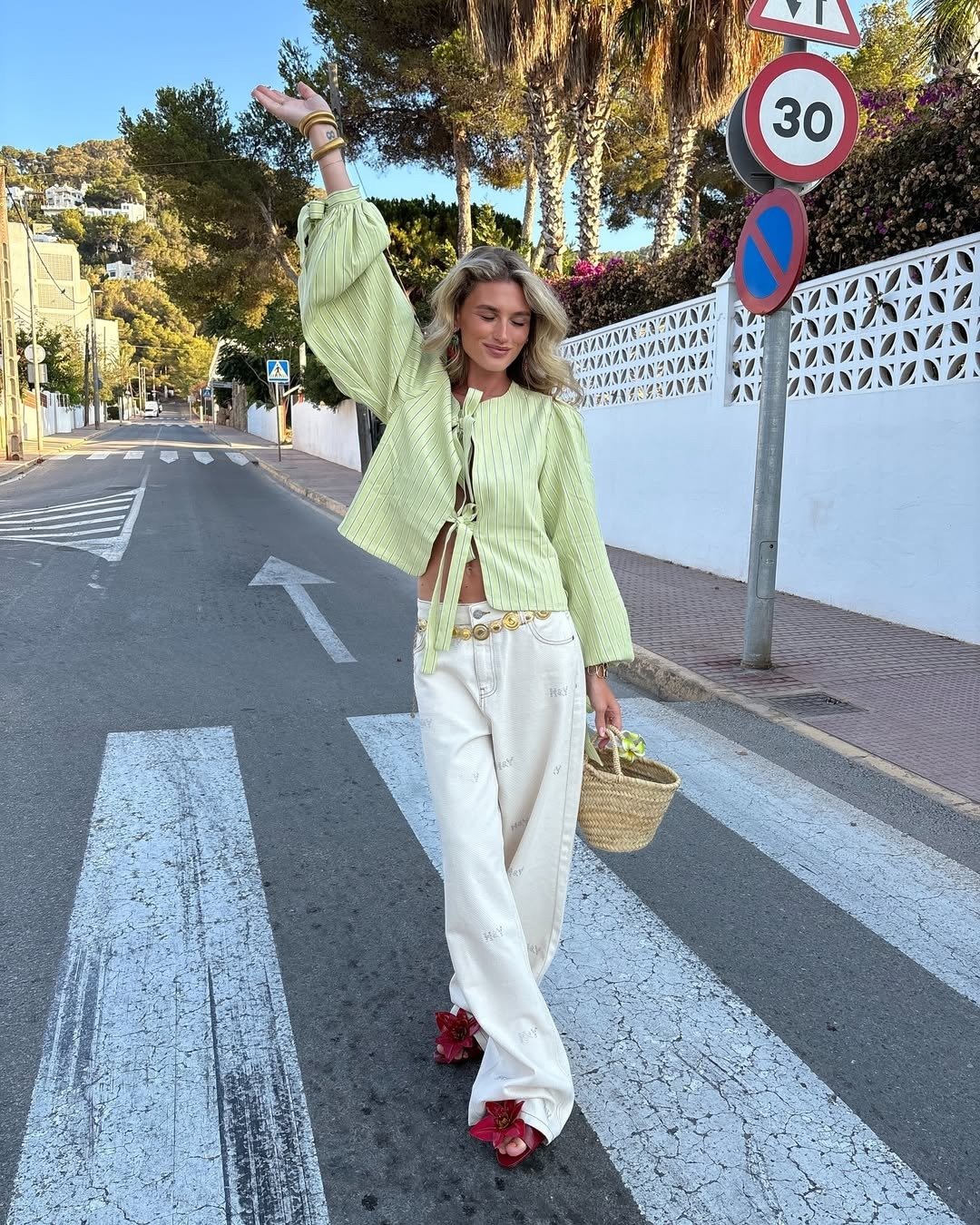 Mulher com blusa verde clara listrada, calça branca ampla e modelos de rasteirinha vermelhos com flor.