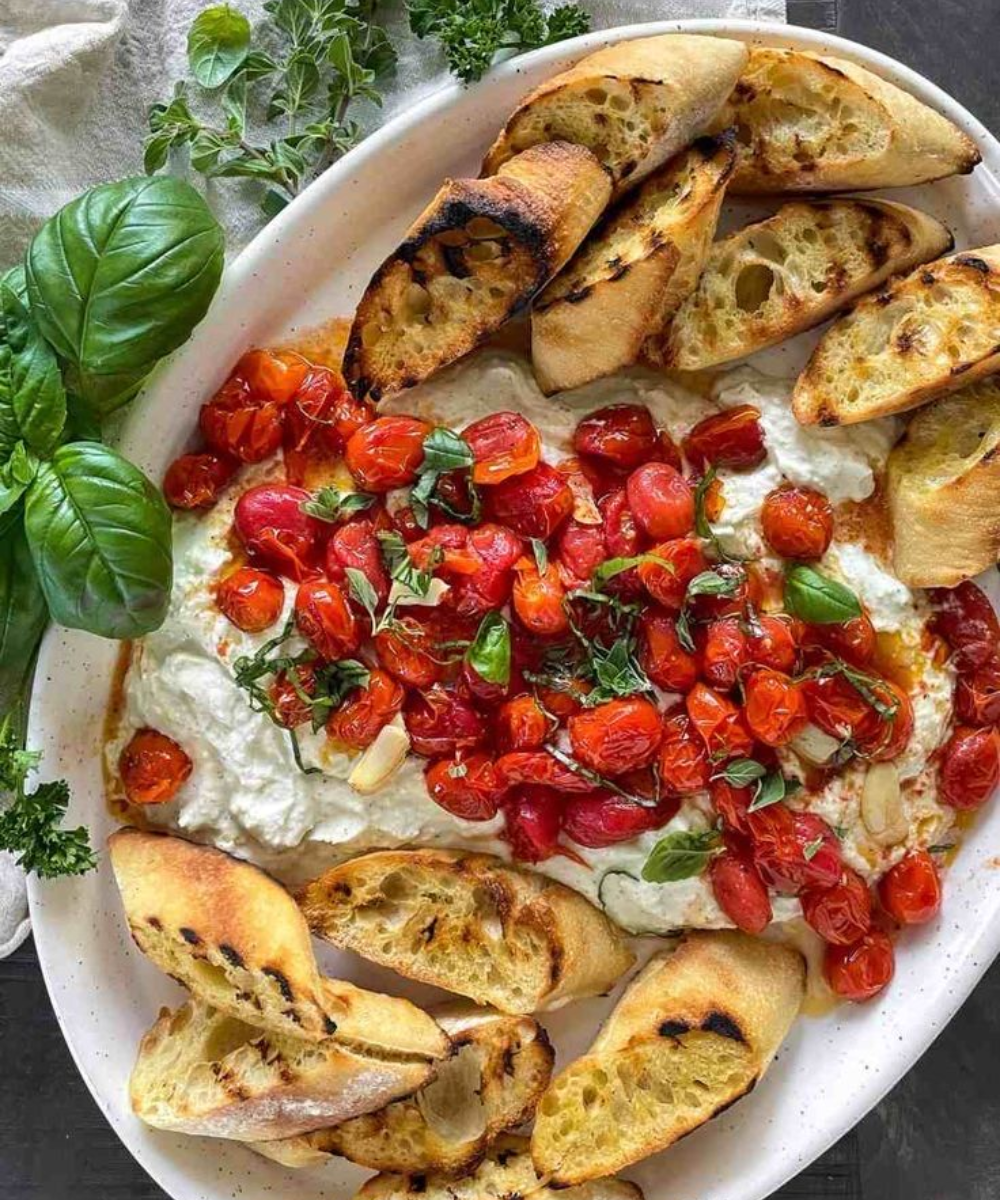 Prato branco com creme de ricota, tomate-cereja assado, folhas de manjericão e fatias de pão artesanal tostado.