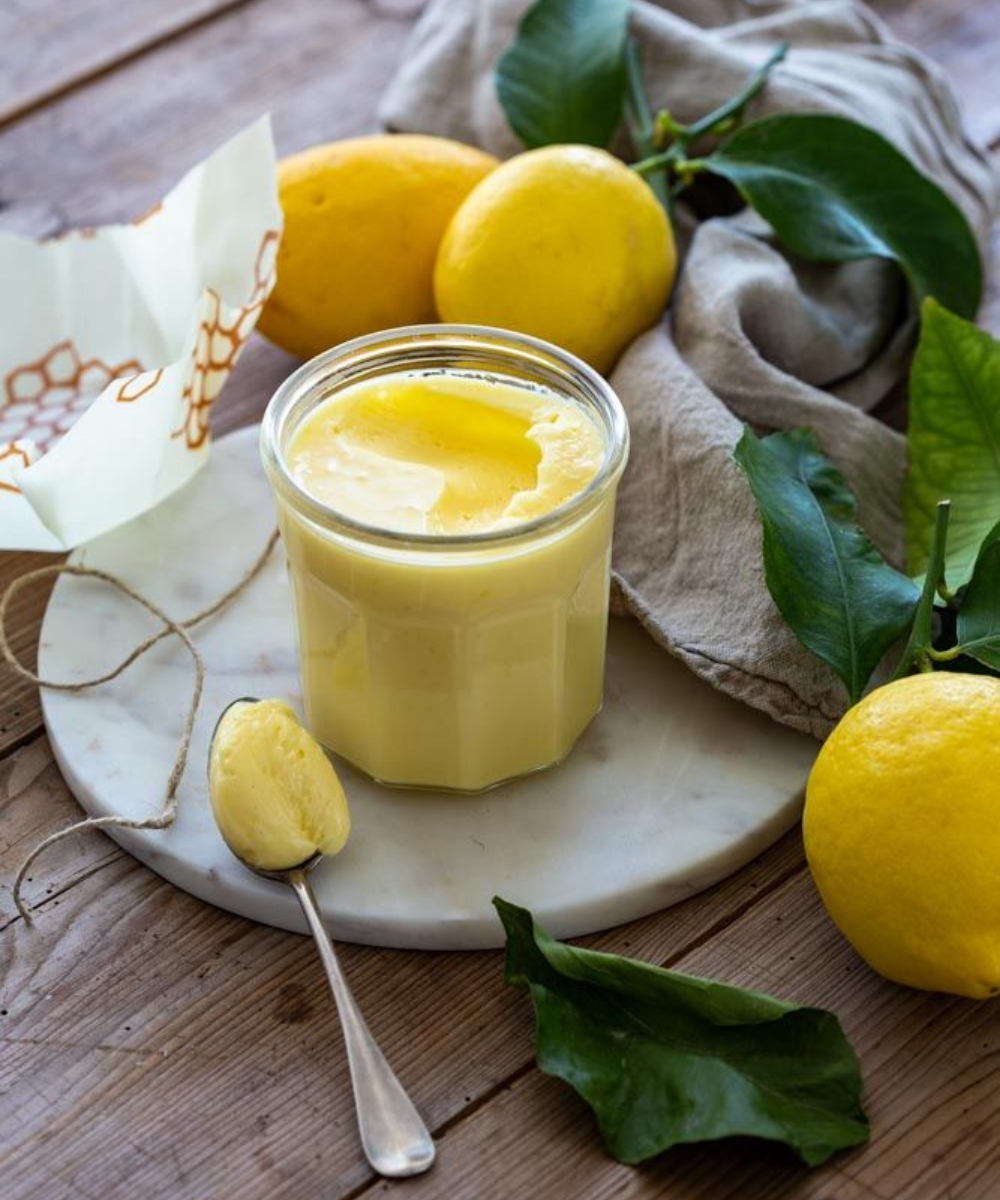 Creme de limão cremoso em pote de vidro sobre base de mármore, cercado por limões amarelos frescos e folhas verdes.