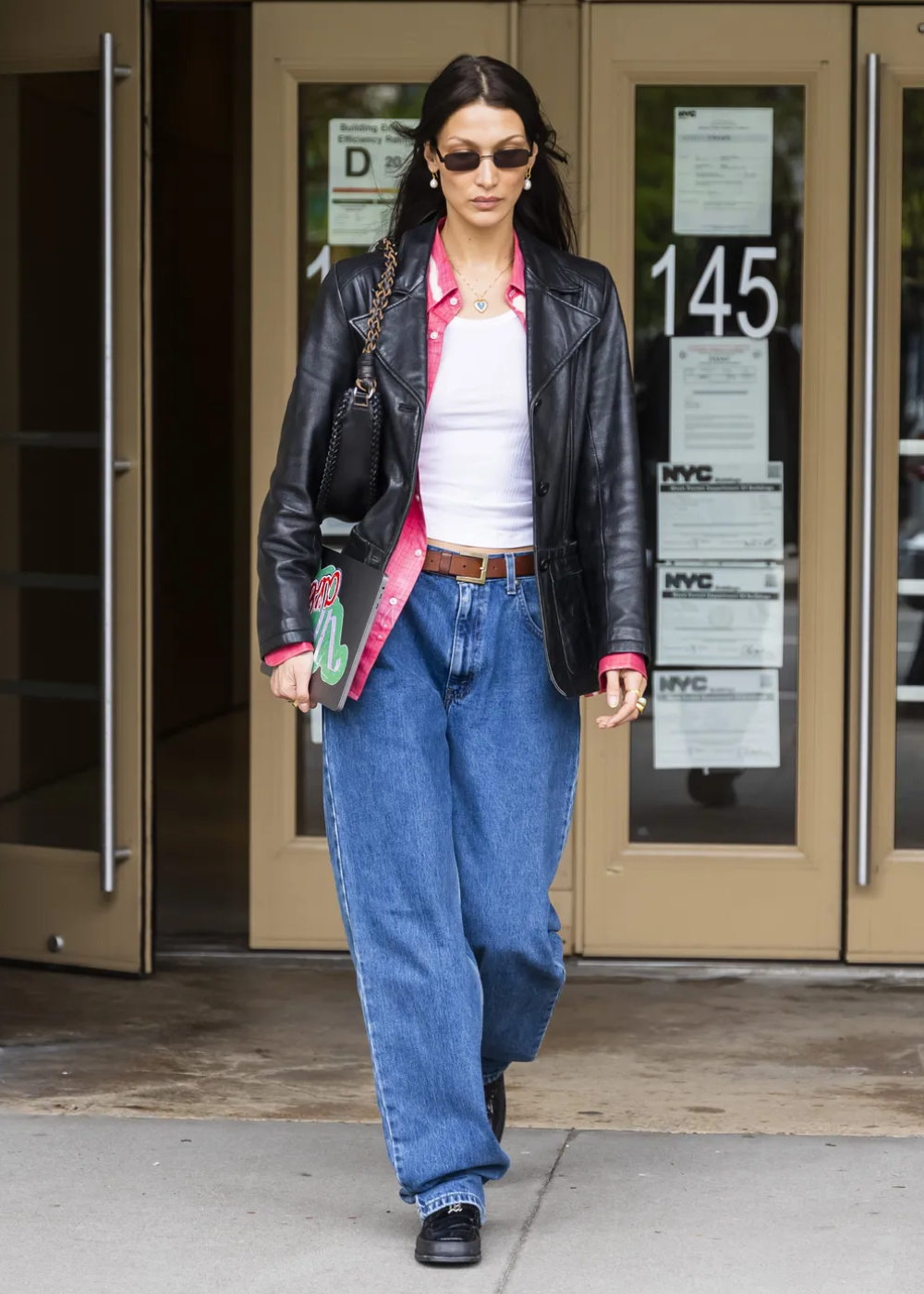 Modelo caminha com regata branca justa e jeans azul de modelagem ampla, combinados com jaqueta de couro preta e camisa rosa.