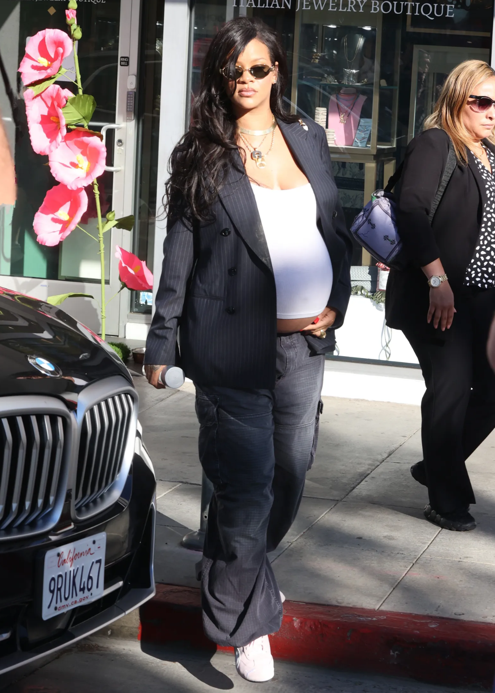 Modelo grávida posa com regata branca justa, blazer preto oversized e jeans cargo cinza, estilo urbano casual e confiante.