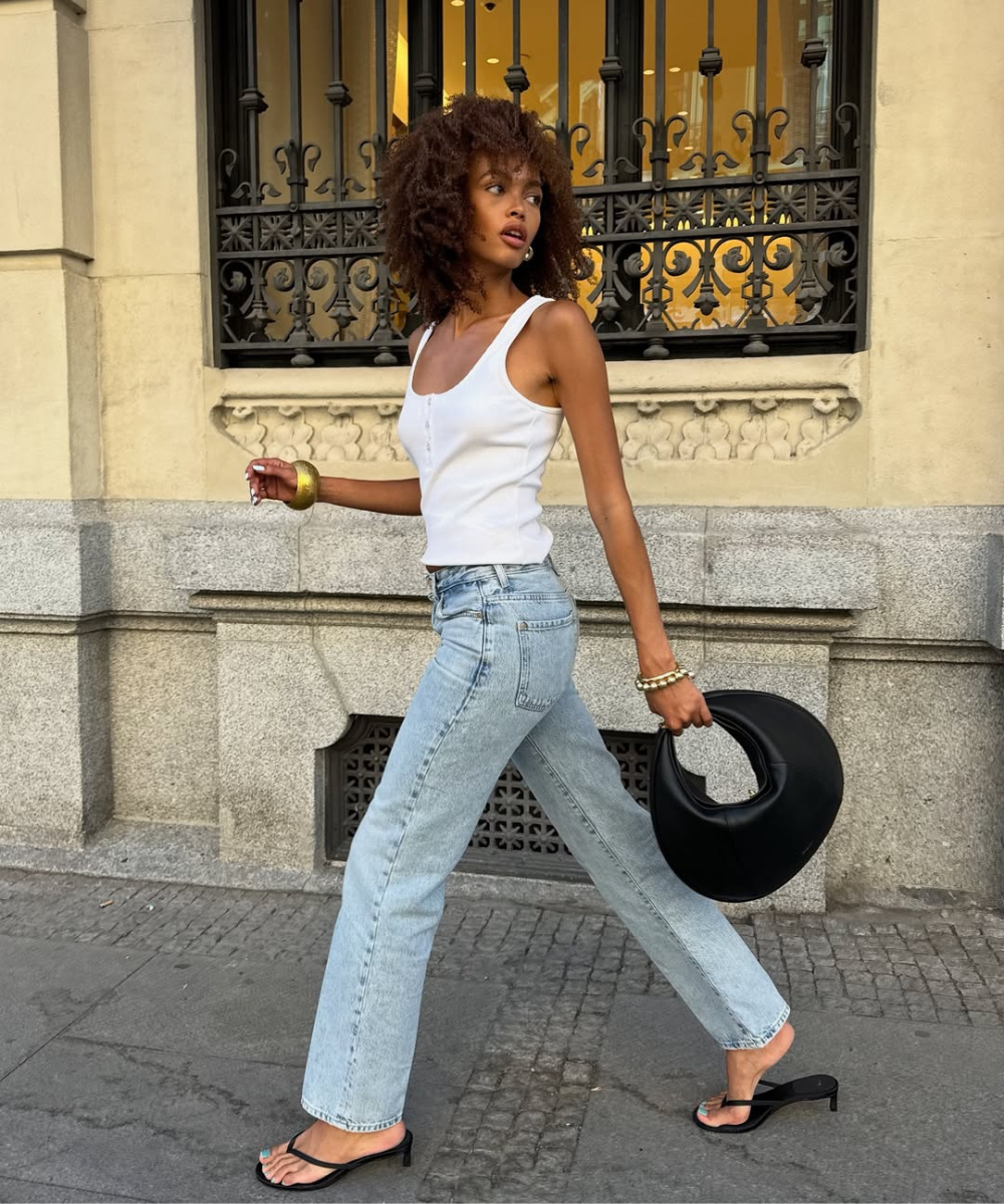 Modelo caminha com postura confiante usando regata branca justa de algodão e jeans claro de corte reto, combinando sandálias