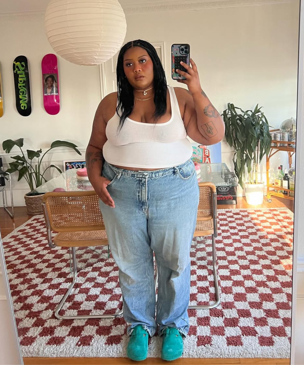 Modelo posa em regata branca justa de malha canelada e jeans azul claro de modelagem ampla, em ambiente casual e acolhedor.