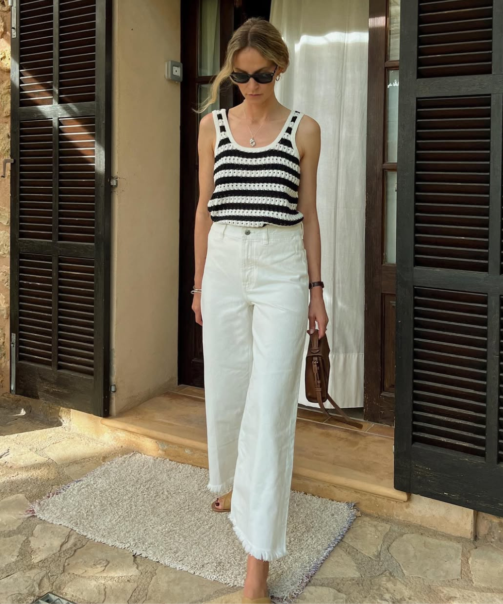 Modelo caminha com regata de crochê listrada preta e branca e jeans branco de barra desfiada, em ambiente rústico.