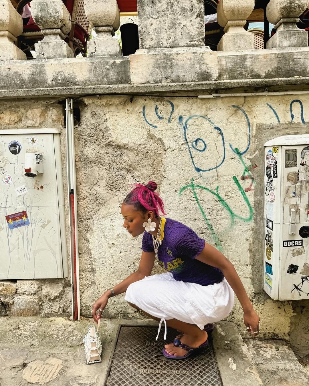 Modelo agacha com postura descontraída usando blusa roxa texturizada, saia branca leve e sandálias roxas