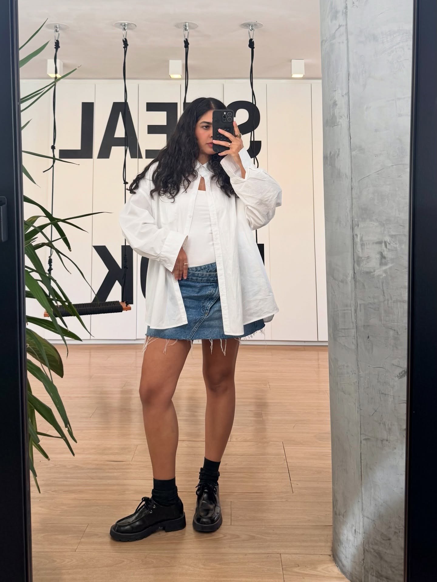 Modelo posa em saia jeans com cós transpassado, camisa branca oversized e sapatos pretos, em ambiente moderno e minimalista.