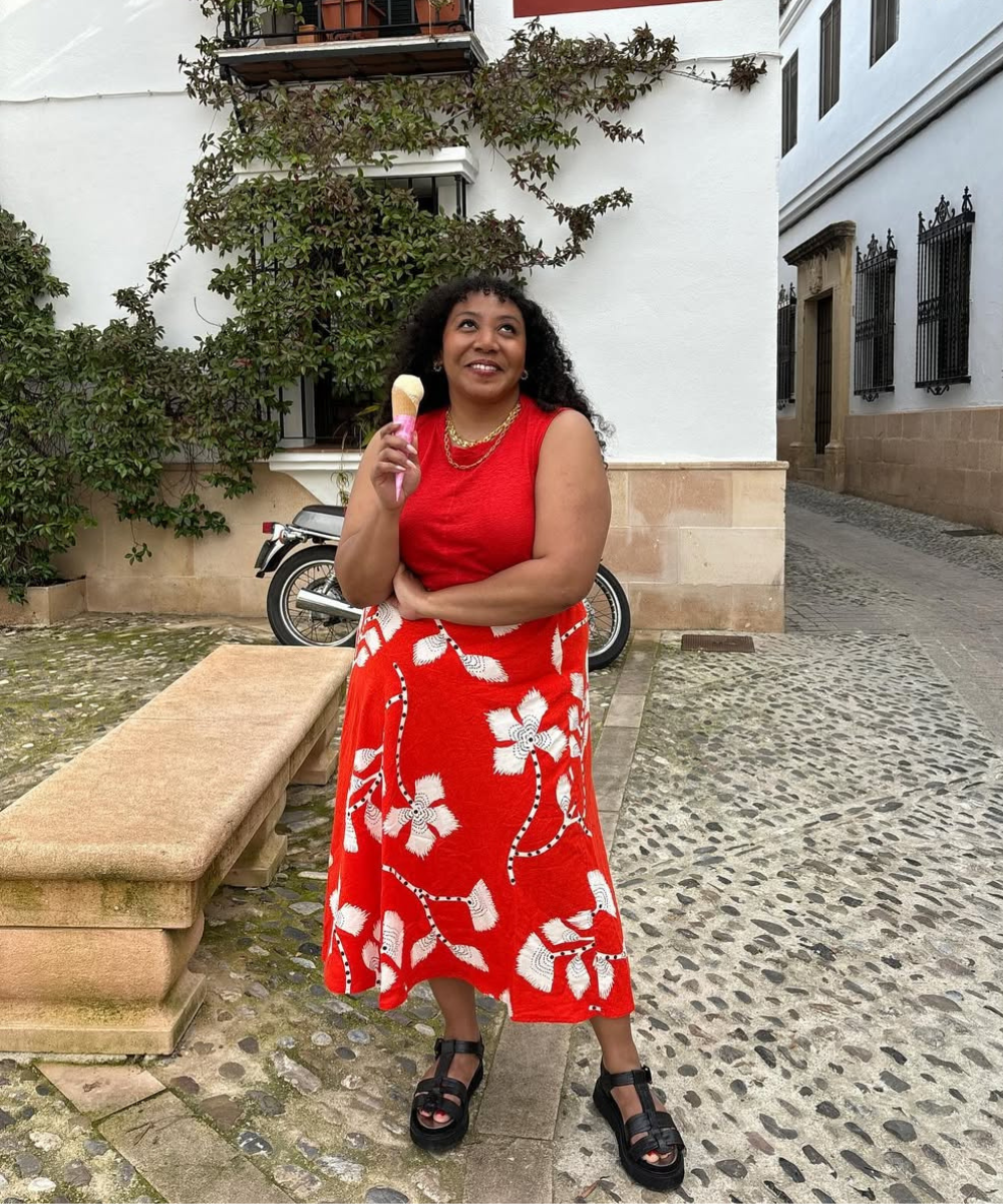 Mulher sorri segurando sorvete, veste saia estampada vermelha com flores brancas e regata vermelha, em rua de pedra.