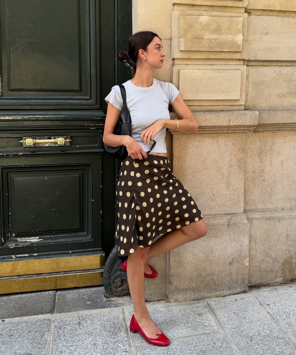 Modelo posa com saia estampada de bolinhas bege sobre fundo marrom em tecido fluido, combinada a camiseta cinza e sapatos