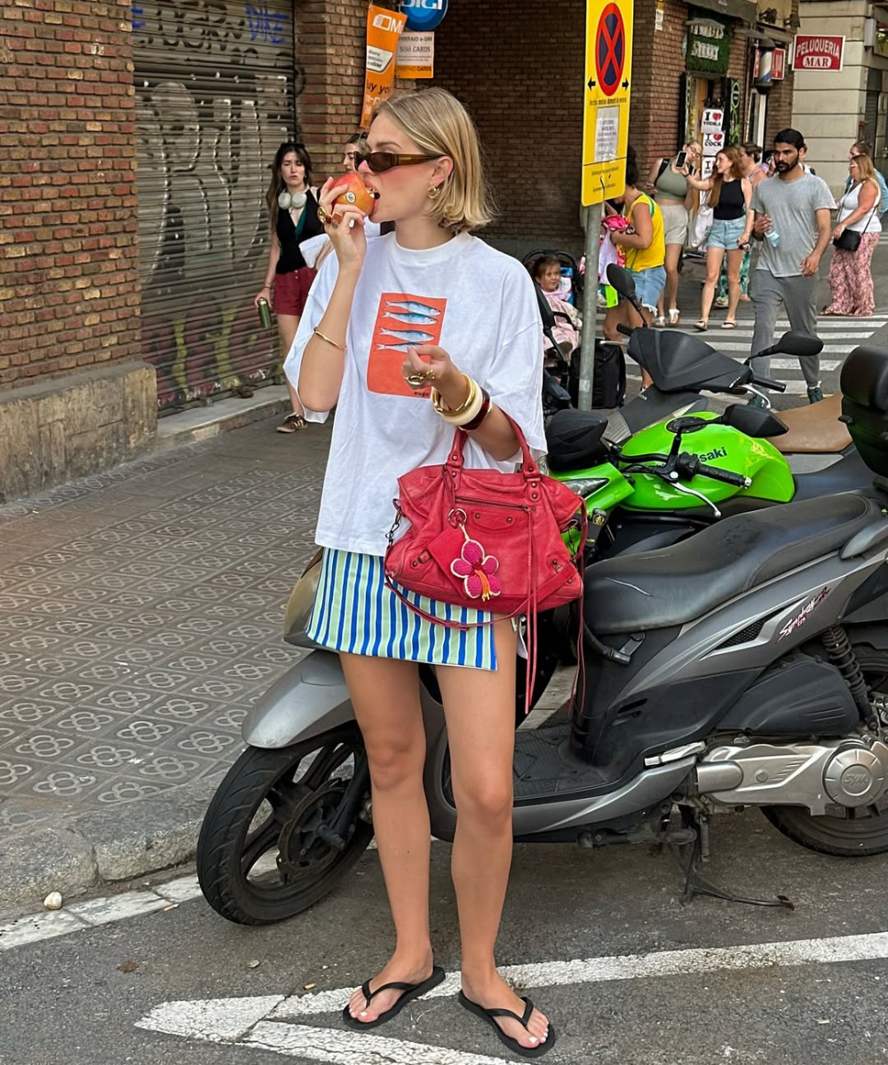 Modelo posa na rua com saia estampada listrada azul e branca, camiseta branca oversized, bolsa vermelha e chinelos pretos.