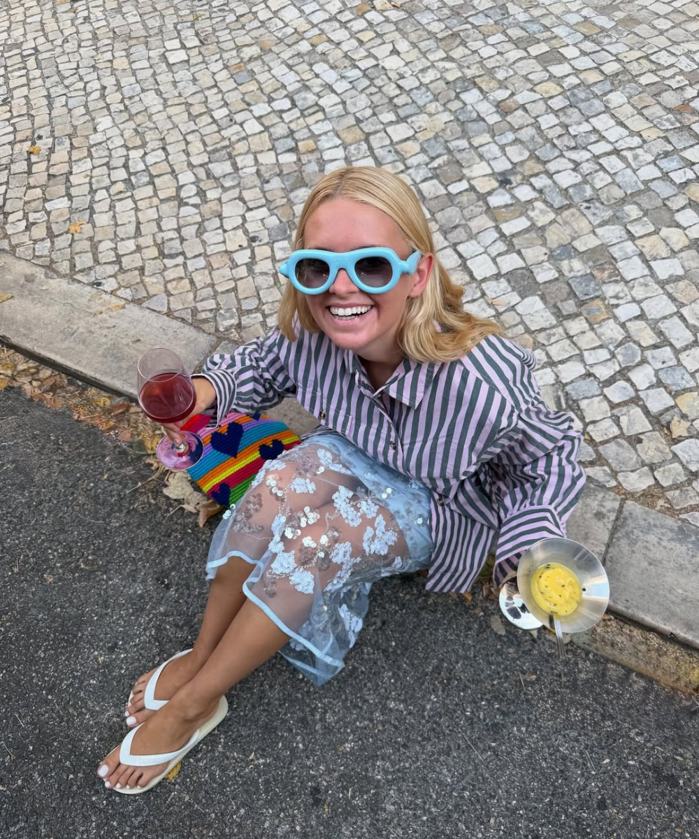 Modelo senta no chão sorrindo, veste saia estampada transparente com flores brancas, blusa listrada e óculos azuis vibrantes.