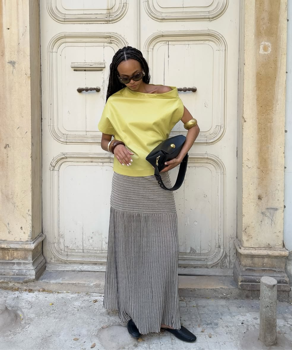 Modelo posa com saia estampada listrada em preto e branco, blusa amarela de tecido estruturado e acessórios dourados.