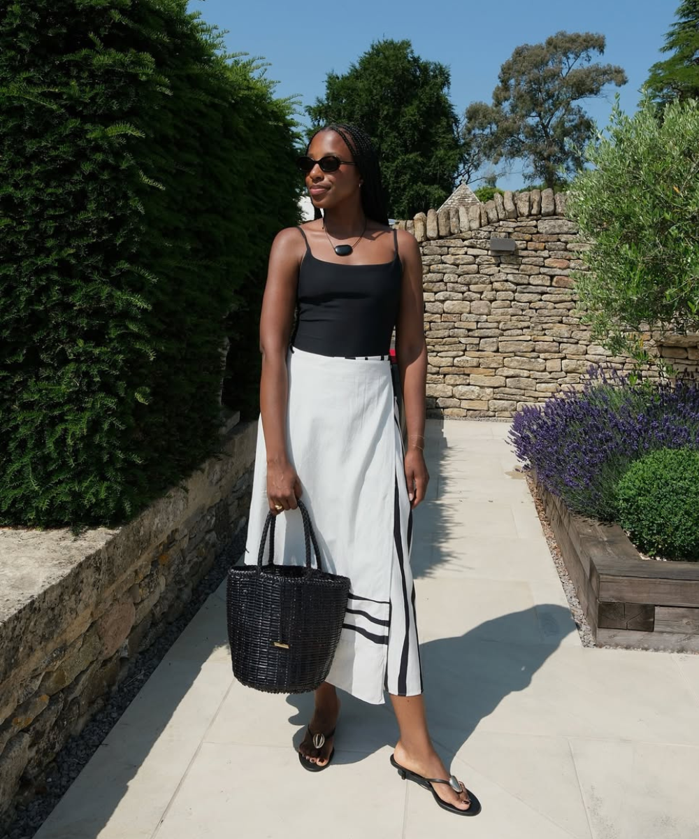 Modelo posa ao ar livre usando saia estampada branca com listras pretas, regata preta e acessórios em preto, clima elegante.