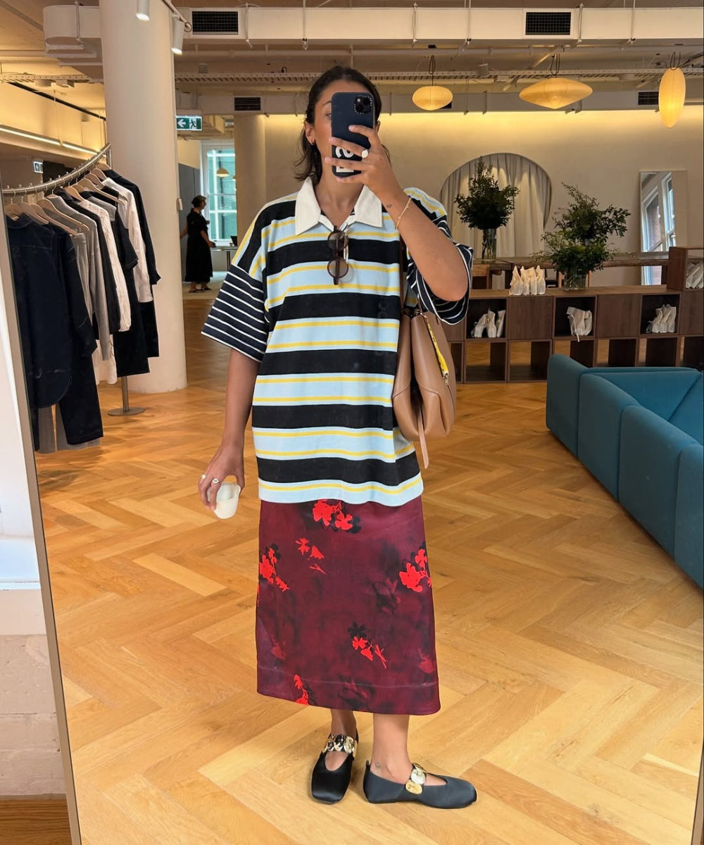 Modelo posa em ambiente interno com saia estampada midi vinho com flores vermelhas, combinada a camiseta listrada oversized.