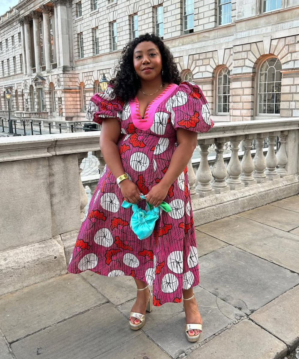 Modelo posa confiante com vestido midi rosa estampado de mangas bufantes e sandália de salto grosso dourada.