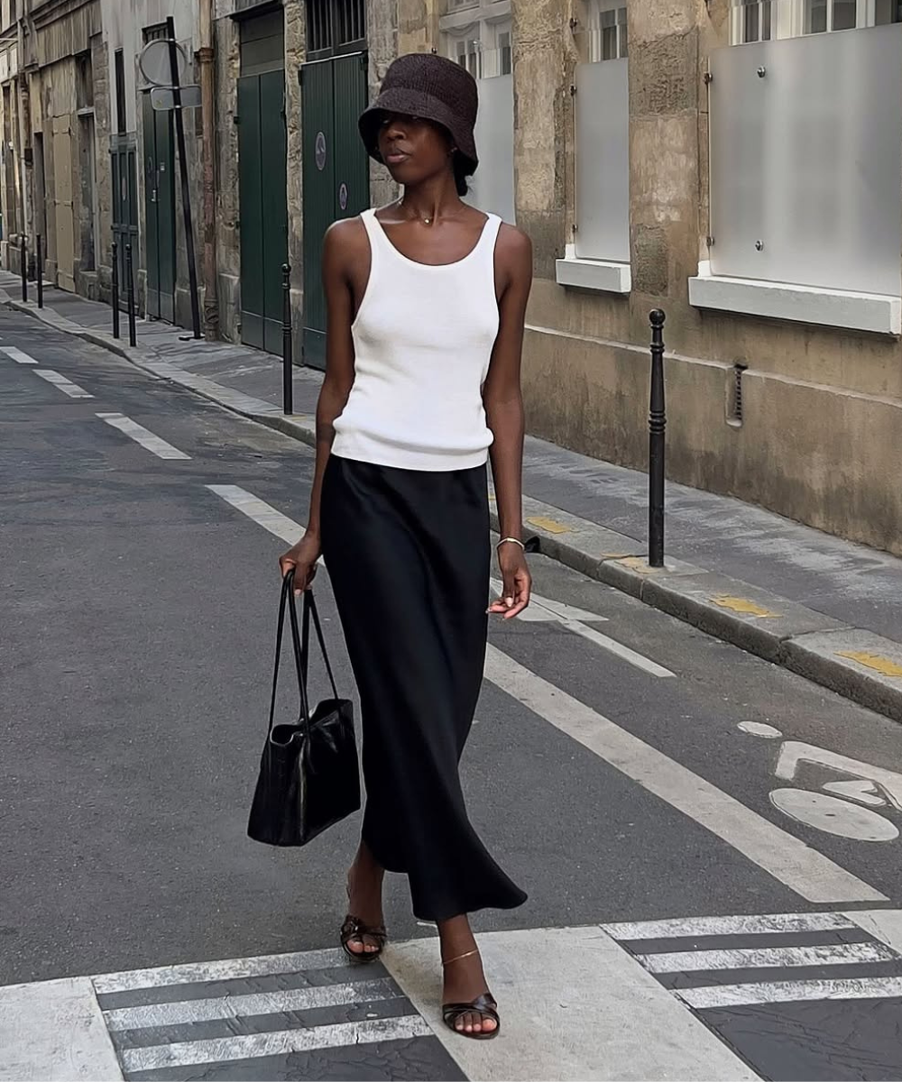 Modelo caminha na rua vestindo regata branca, saia preta fluida, chapéu bucket marrom e sandálias de verão de tiras finas.