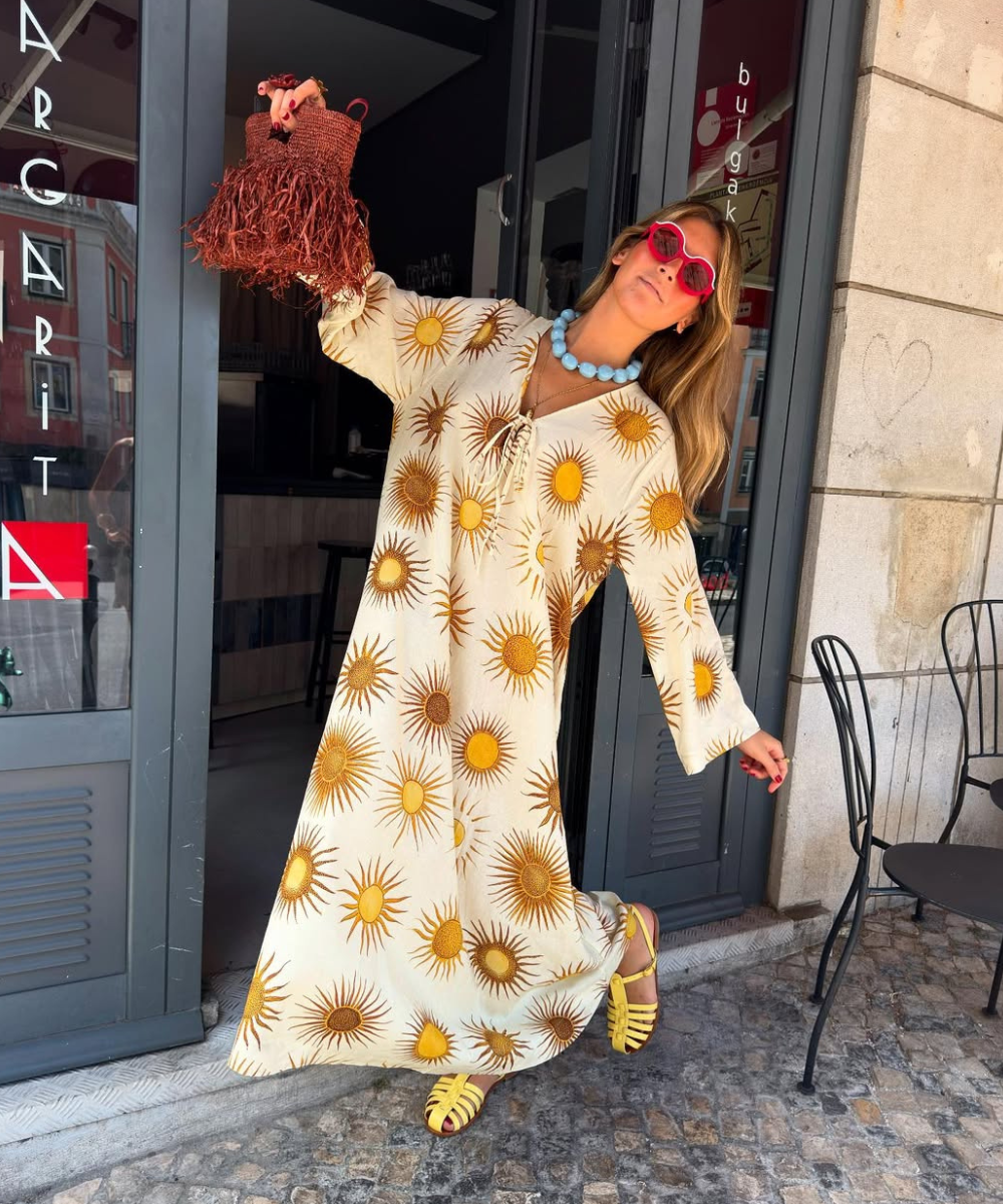 Modelo posa com vestido longo branco estampado de sóis amarelos, sandálias de verão amarelas em tiras e acessórios.