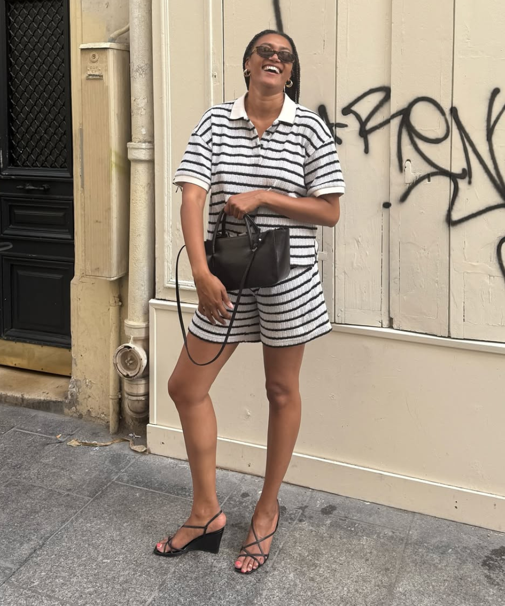 Modelo sorri e posa com sandálias de verão pretas de tiras finas e salto anabela, combinando short e camisa listrados.